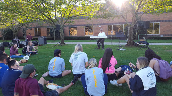 Students sitting down outside listening to musician.