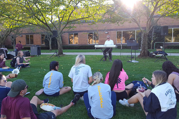 Students sitting down outside listening to musician.